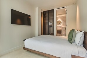 Un dormitorio con cama, televisor montado en la pared y baño visible a través de una puerta abierta.