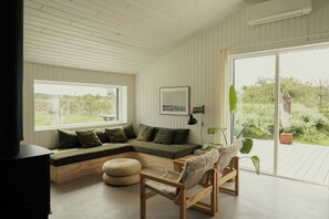 A modern living room with a sectional sofa, a wooden coffee table, a small round ottoman, and a wooden chair. There is a large window providing a view of greenery outside. - Nordic cottage by Nationalpark Thy (Klitmøller)