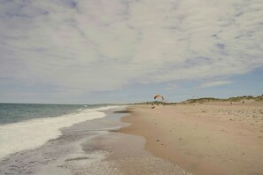 Uma praia com ondas, uma duna e uma pessoa empinando pipa.