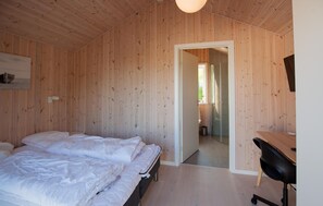 Quarto com paredes de madeira, uma cama com roupa de cama branca, uma escrivaninha com uma cadeira e uma porta que dá para outro espaço.