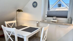 Ein Zimmer mit weißem Tisch und Stühlen, einem Heizkörper, einem Fenster mit Blick auf ein Gebäude und einem dekorativen Teller an der Wand.