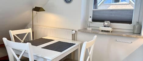 Una habitación con una mesa y sillas blancas, un radiador, un ventanal con vistas a un edificio y un plato decorativo en la pared.