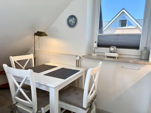Ein Zimmer mit weißem Tisch und Stühlen, einem Heizkörper, einem Fenster mit Blick auf ein Gebäude und einem dekorativen Teller an der Wand.