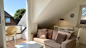 Ein gemütliches Wohnzimmer mit einer Couch, einem kleinen Tisch und einem Sessel. Ein Fenster bietet Blick auf ein Haus und Bäume.