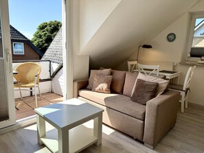 Ein gemütliches Wohnzimmer mit einer Couch, einem kleinen Tisch und einem Sessel. Ein Fenster bietet Blick auf ein Haus und Bäume.