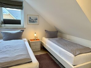 A bedroom with two single beds, a nightstand with a lamp, a window with blinds, and a framed picture on the wall. - Reiherweg, Apartment 3 - Beautiful 2-room vacation apartment with a south-facing balcony, quiet and (Sylt)