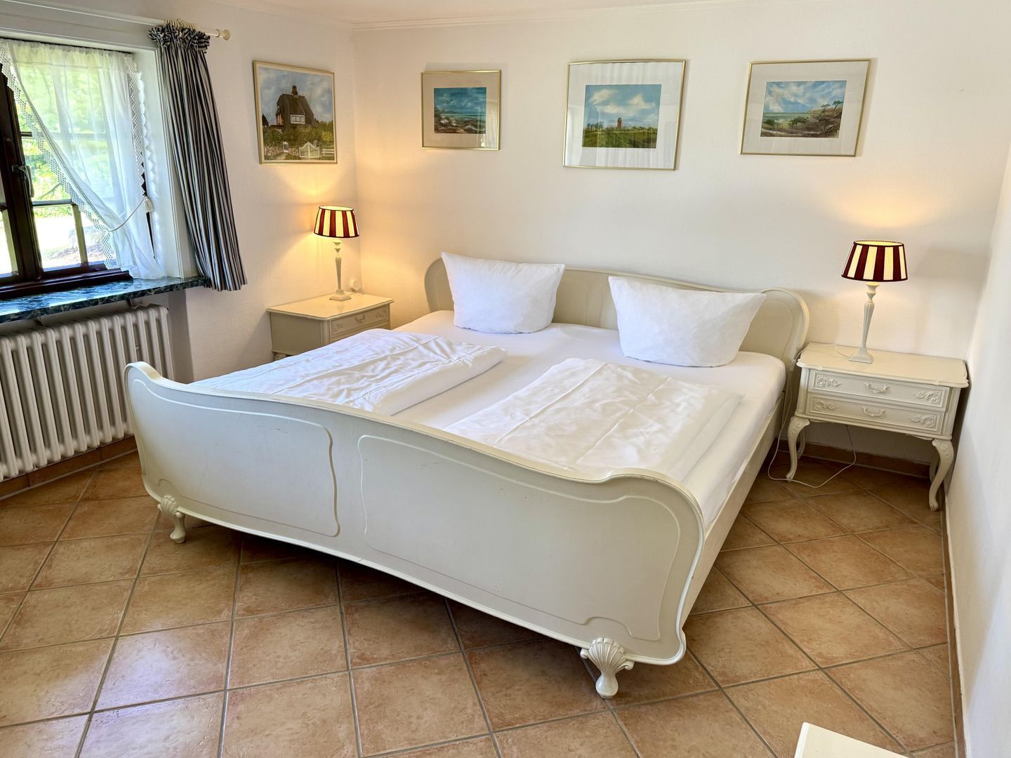 A bedroom with a bed, two bedside tables, a window with curtains, and three framed pictures on the wall.