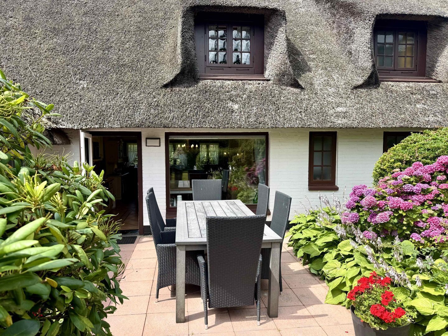Een vakantiehuis met rieten dak, een patio tafel en stoelen, omgeven door planten en bloemen.