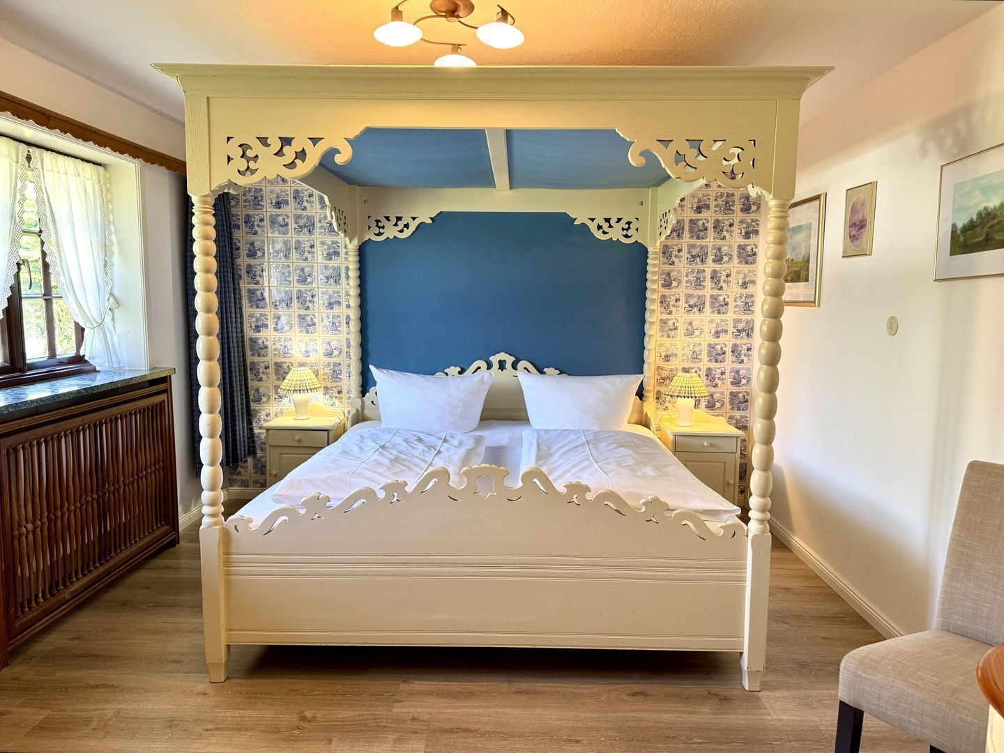 Una cama con dosel sobre un piso de madera, en una habitación con papel tapiz estampado. La cama tiene un cabecero azul y está flanqueada por dos mesitas de noche con lámparas.