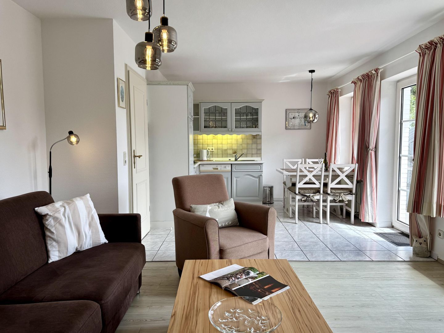 Un salon moderne avec un canapé marron, une table basse en bois sur laquelle sont posés des magazines, et un coin repas avec des chaises blanches.
