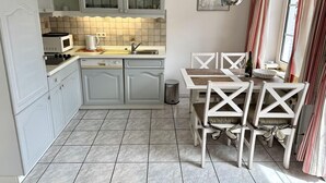 A compact kitchen with white cabinetry, a microwave, and a sink. There is a dining table with chairs and a window with curtains. - Süderstraße, Apartment 05 – Landhaus - Cozy 2-room vacation apartment for 4 (Sylt)
