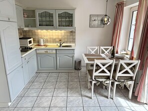 A compact kitchen with white cabinetry, a microwave, and a sink. There is a dining table with chairs and a window with curtains. - Süderstraße, Apartment 05 – Landhaus - Cozy 2-room vacation apartment for 4 (Sylt)