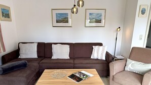 A living room with a brown sectional sofa, a wooden coffee table, and a pink armchair. There are framed pictures on the wall and a floor lamp. - Süderstraße, Apartment 05 – Landhaus - Cozy 2-room vacation apartment for 4 (Sylt)
