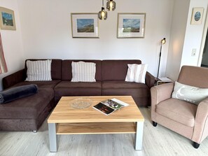 A living room with a brown sectional sofa, a wooden coffee table, and a pink armchair. There are framed pictures on the wall and a floor lamp. - Süderstraße, Apartment 05 – Landhaus - Cozy 2-room vacation apartment for 4 (Sylt)