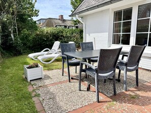 Een terras buiten met een tafel en stoelen, omringd door een tuin en een wit gebouw.