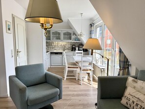 Eine moderne Küche mit weißen Schränken und einem Essbereich mit Tisch und Stühlen. Im Vordergrund befindet sich eine Couch mit Kissen und eine Lampe.