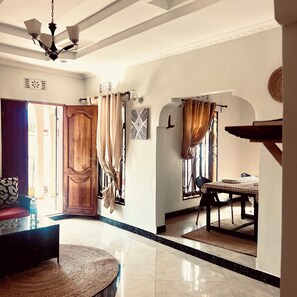 A hotel lobby with a wooden door, a round woven rug, a dining area with a table and chairs, and a ceiling fan.