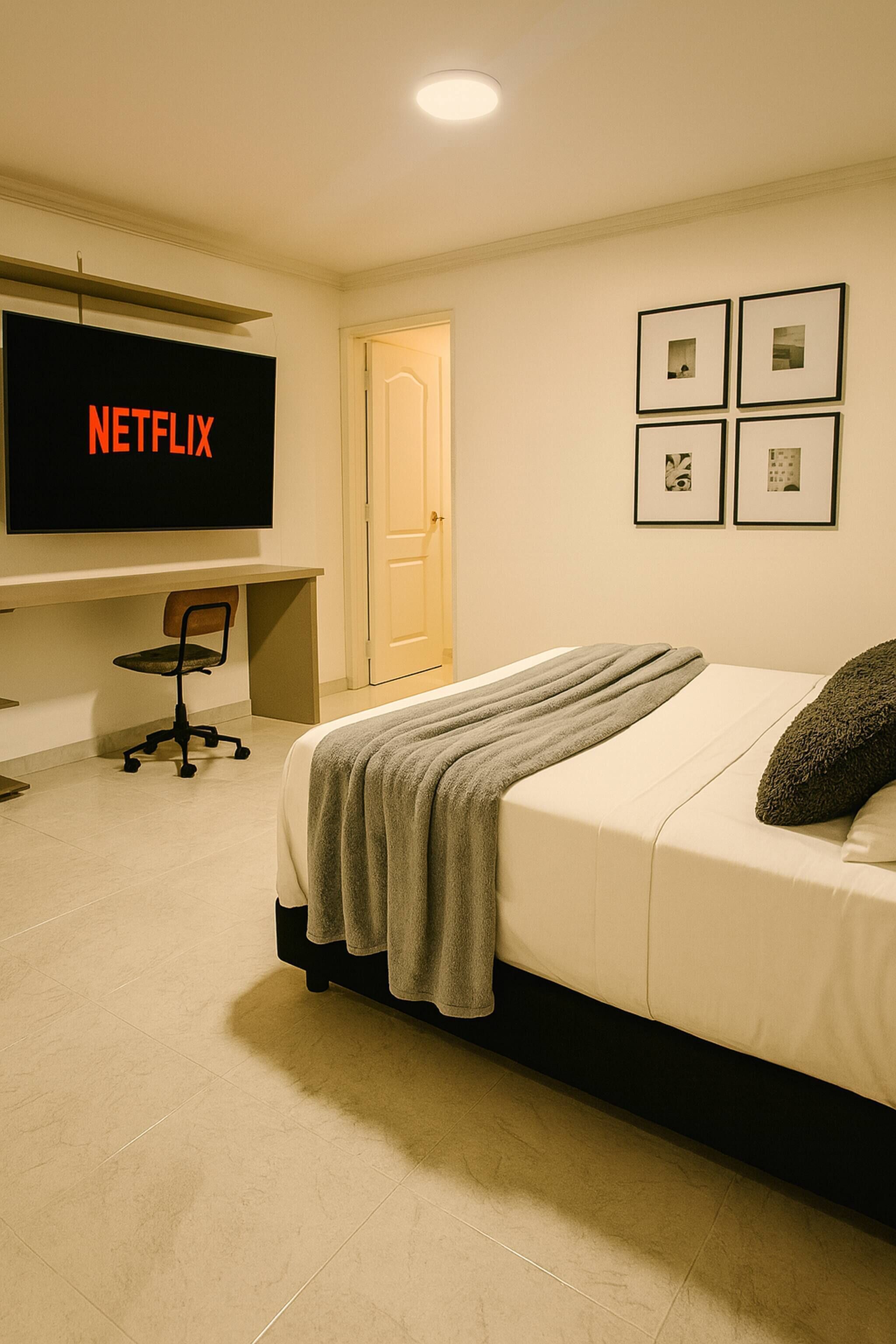 A bedroom with a bed, a desk with a computer, a chair, and a television displaying the Netflix logo.