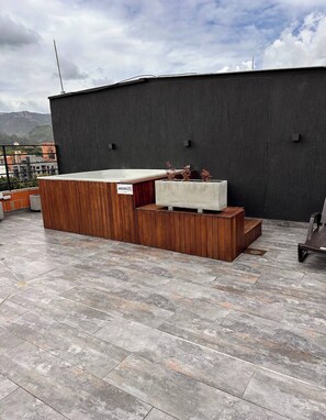 Een dakterras met een houten bankje, een betonnen plantenbak met planten en een metalen barbecue.
