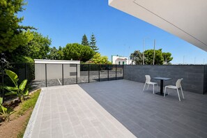 Een terras met een tafel en stoelen, een klein afgesloten gebouwtje, en een uitzicht op de omliggende groenvoorziening en gebouwen.