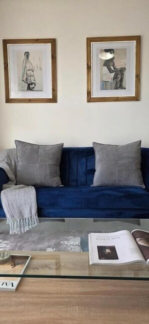 A room with a blue sofa, two framed artworks, a glass coffee table with a magazine, and a decorative throw.