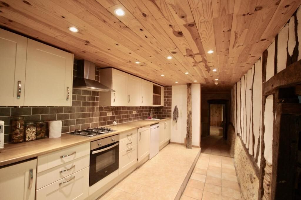 Une cuisine avec des poutres apparentes en bois au plafond, des meubles blancs, une cuisinière à gaz et un dosseret carrelé.