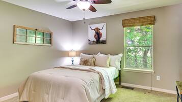 A bedroom with a bed, a ceiling fan, a window with a view of trees, and a wall-mounted picture of a cow.