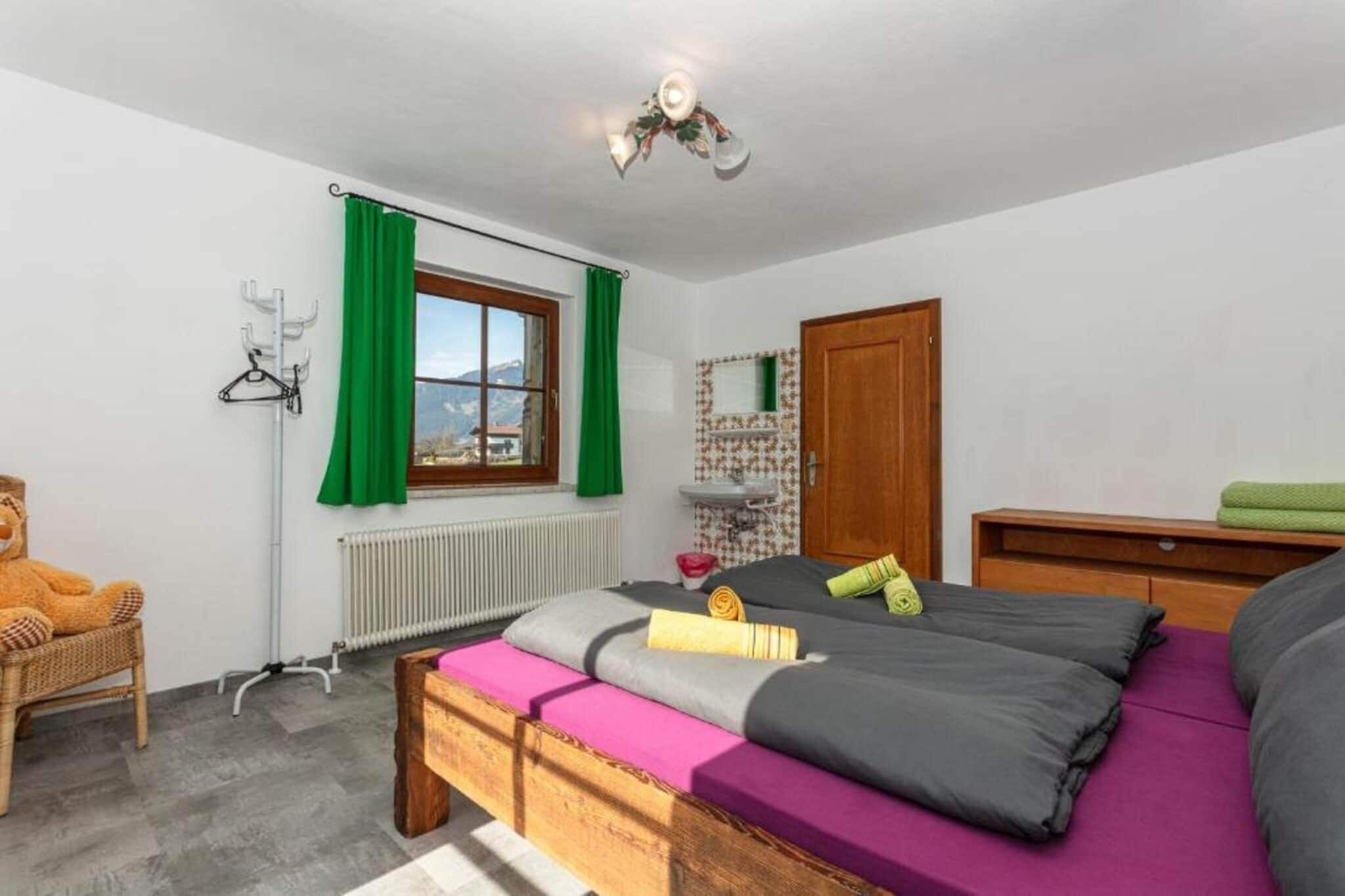 A bedroom with a bed, a chair, a radiator, a window with curtains, a wooden door, and a small shelf with a mirror.
