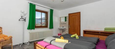 Un dormitorio con cama, una silla, un radiador, una ventana con cortinas, una puerta de madera y un estante pequeño con un espejo.