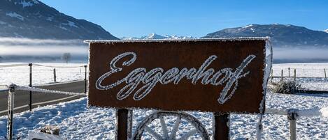 Un letrero con el nombre 'Eggenhof' en cursiva, montado en un poste metálico con una rueda, frente a un paisaje nevado con montañas al fondo.