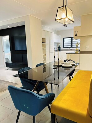 A modern dining area with a glass table, blue chairs, and a yellow bench. - Luxurious 3-Bedroom Apartment in Johannesburg's Waterfall Area (Johannesburg)