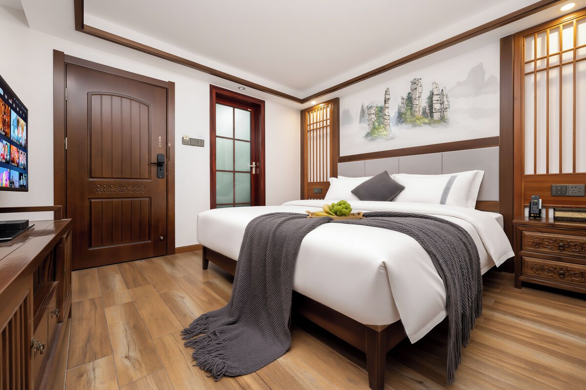 Quarto de hotel com piso de madeira, cama com roupa de cama branca, manta cinza e televisão fixada na parede.