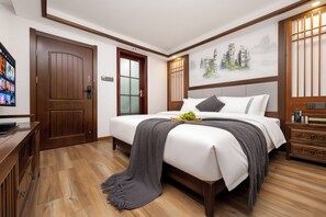 Habitación de hotel con piso de madera, cama con ropa blanca, una manta gris y un televisor montado en la pared.