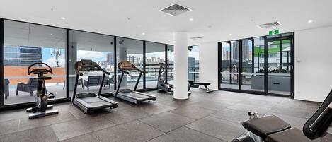 A modern gym with cardio machines, weight benches, and large windows offering city views.