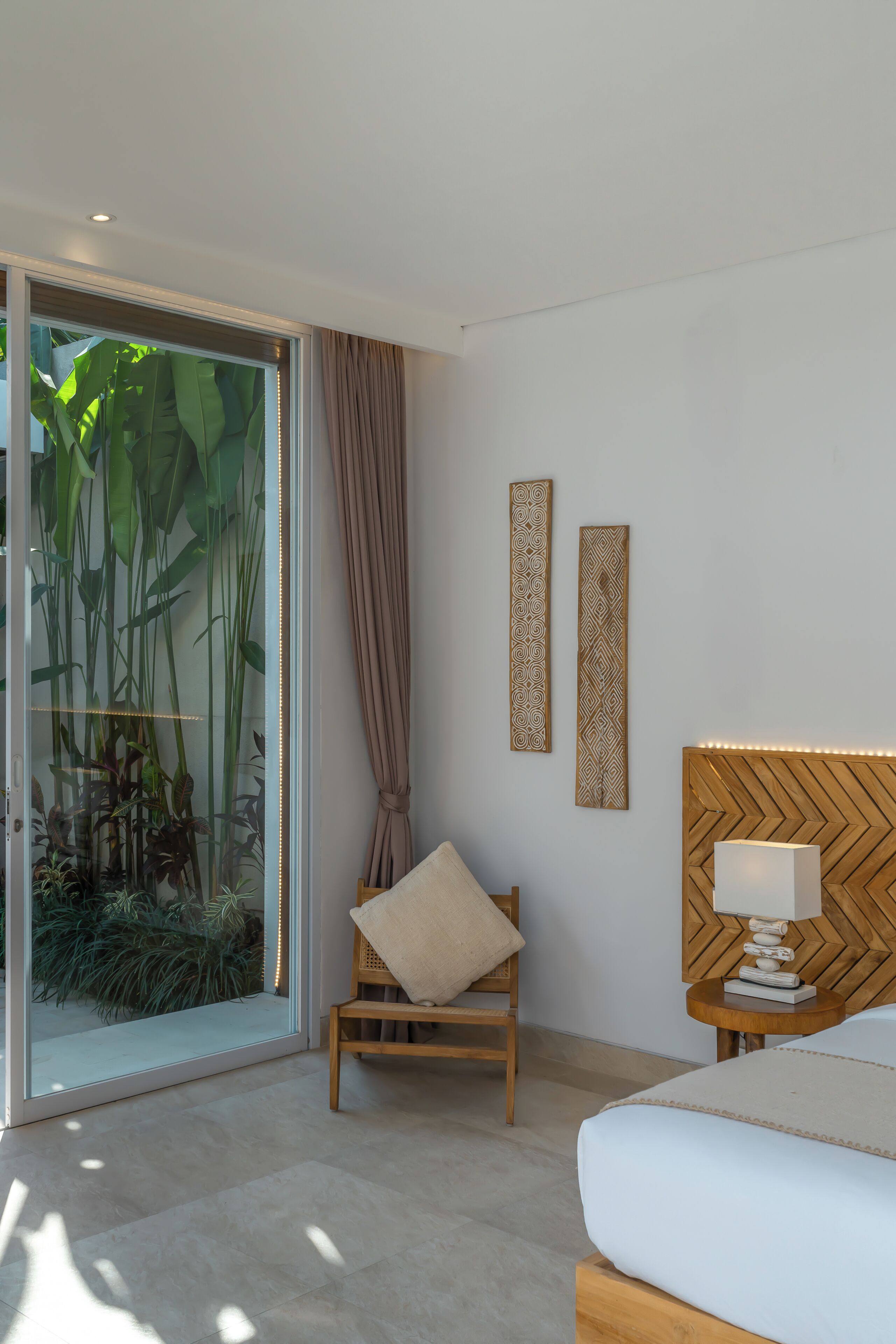 A modern bedroom with a glass door leading to a garden, a wooden chair, a bedside table with a lamp, and two decorative panels on the wall.