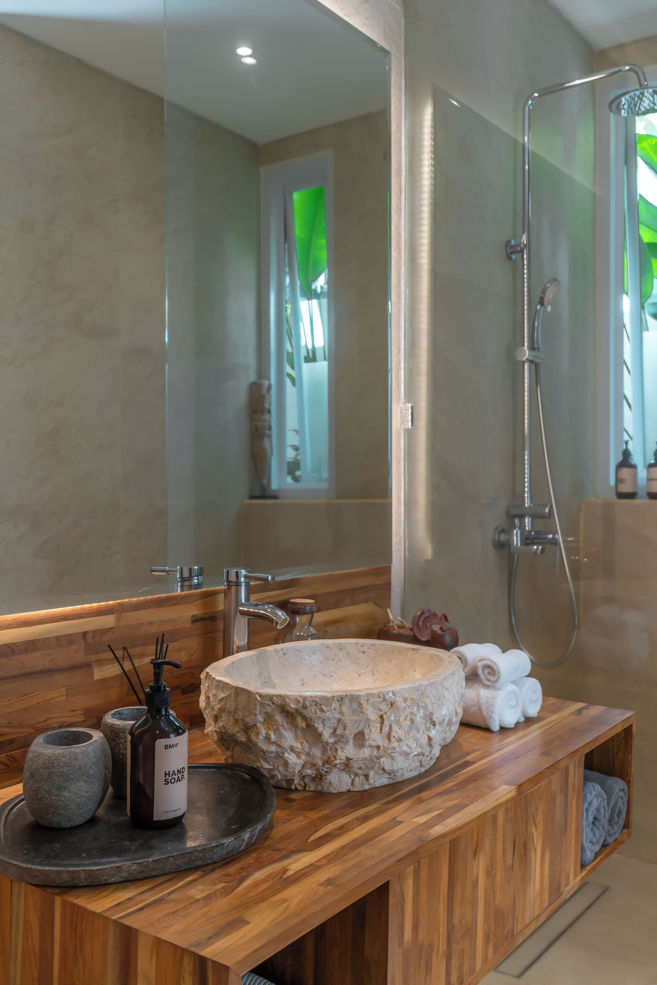 Un baño moderno con tocador de madera, lavamanos de piedra, dispensador de jabón manual y cabina de ducha de vidrio.