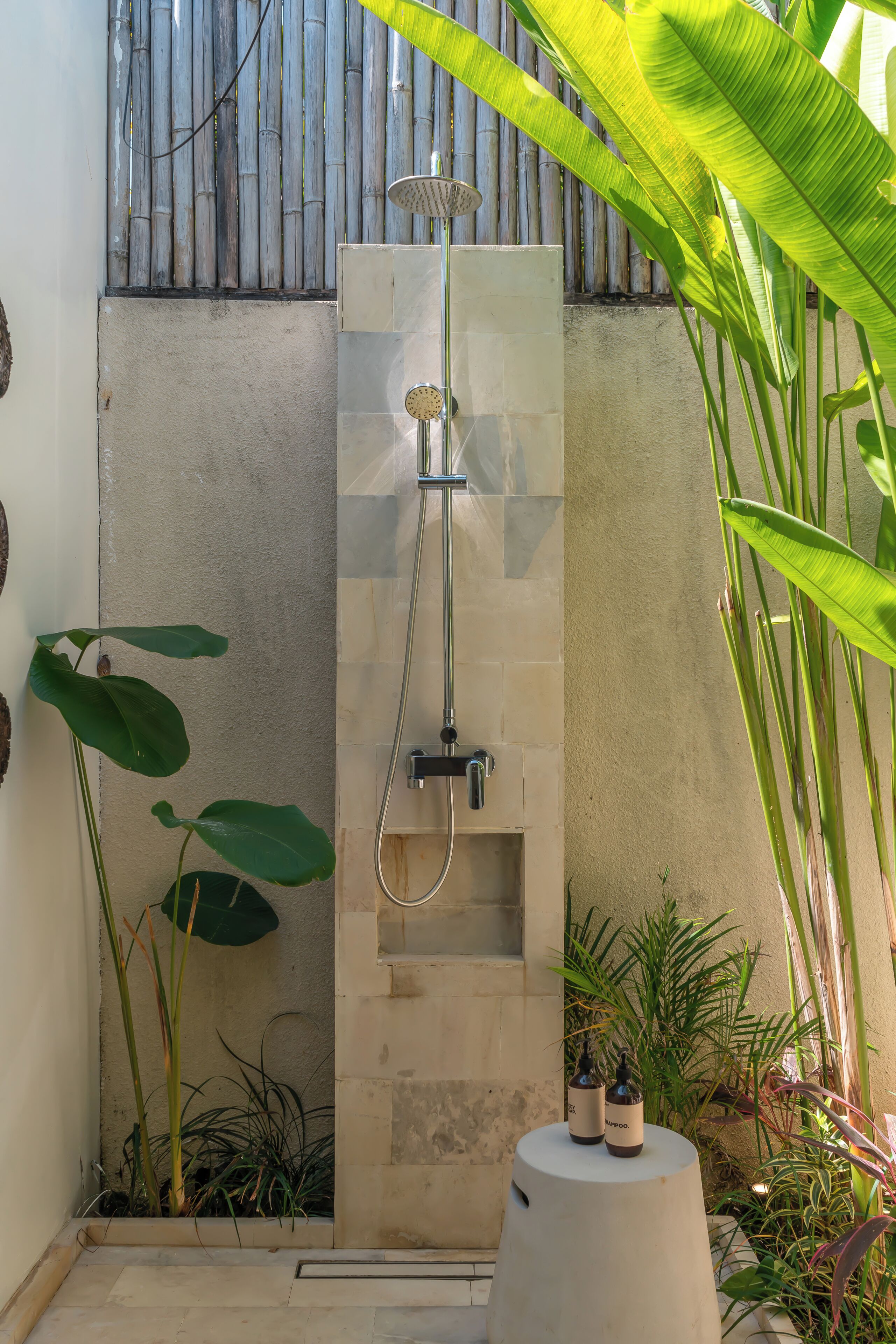Zona de ducha con cabezal de ducha tipo lluvia vertical, un escalón para acceder y un pedestal blanco con botellas de champú.