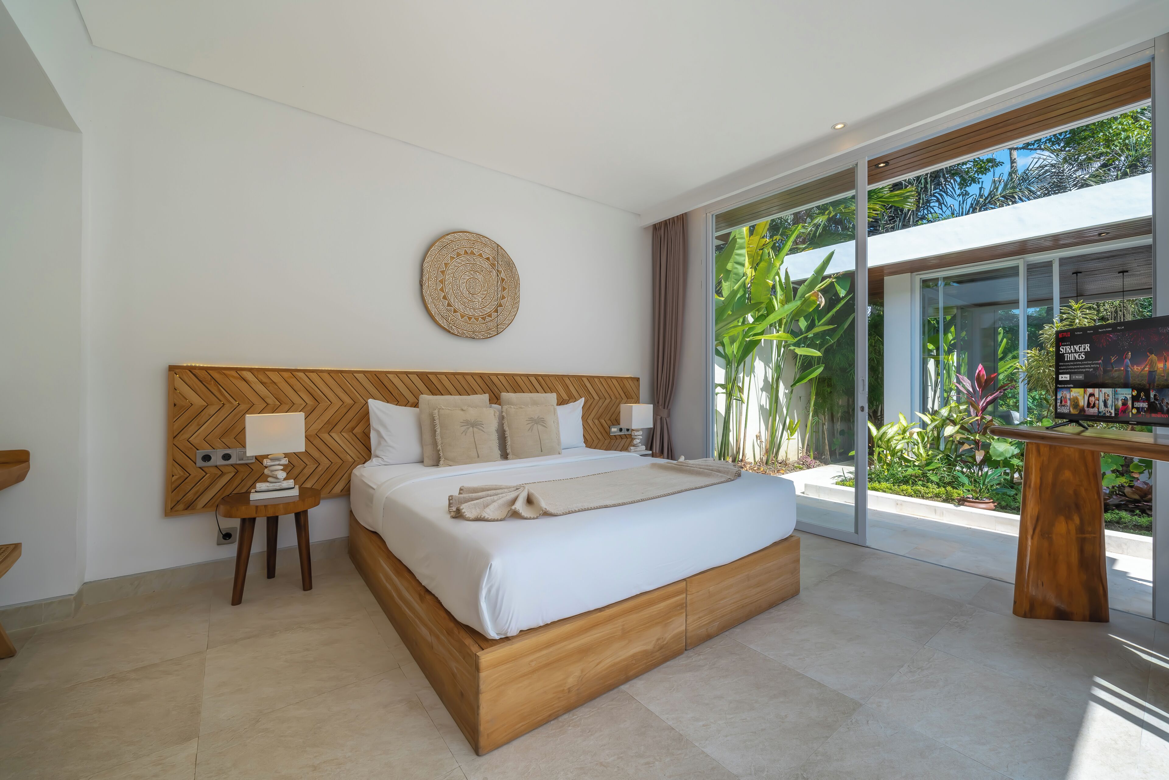 Una habitación de hotel moderna con una cama grande, cabecera de madera, mesita de noche con lámpara y vistas al exterior a través de amplias ventanas.