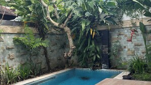 En swimmingpool med et træbord i forgrunden, omgivet af frodig vegetation og en stenvæg.