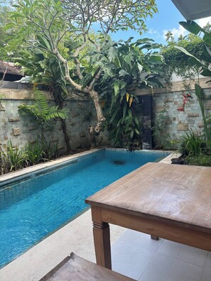 A swimming pool with a wooden table in the foreground, surrounded by lush greenery and a stone wall. - 3 BR, 250 Sqmt villa, Terrace, 10 min Finns (Kecamatan Kuta Utara)