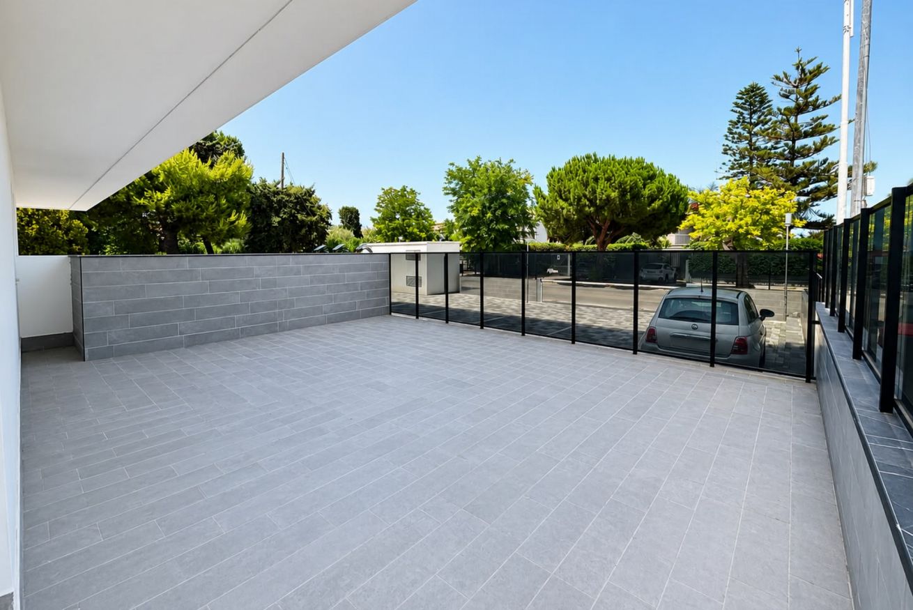 Un espace extérieur abrité, doté d’un sol carrelé, avec une voiture garée au fond et une vue dégagée sur des arbres et un bâtiment situé de l’autre côté de la rue.