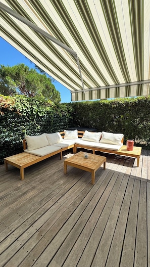 Un espacio de descanso al aire libre con techado, que cuenta con una terraza de madera, un conjunto de sofás y una mesa de centro.