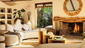 A cozy living room with a fireplace, a sofa, a coffee table, and a bookshelf with decorative items. - Cottar's Private Bush Villa (Ololaimutiek Village)