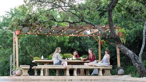 A group of people seated at a wooden table under a pergola with string lights, surrounded by greenery and a pool. - Cottar's Private Bush Villa (Ololaimutiek Village)