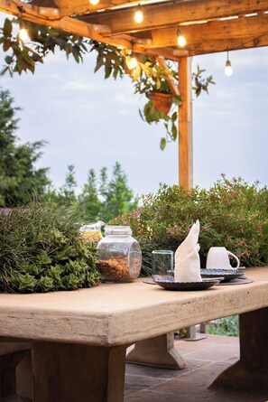 Un área de comedor al aire libre con una pérgola de madera, luces colgantes, una mesa dispuesta con platos, un tarro de vidrio y un paño blanco.
