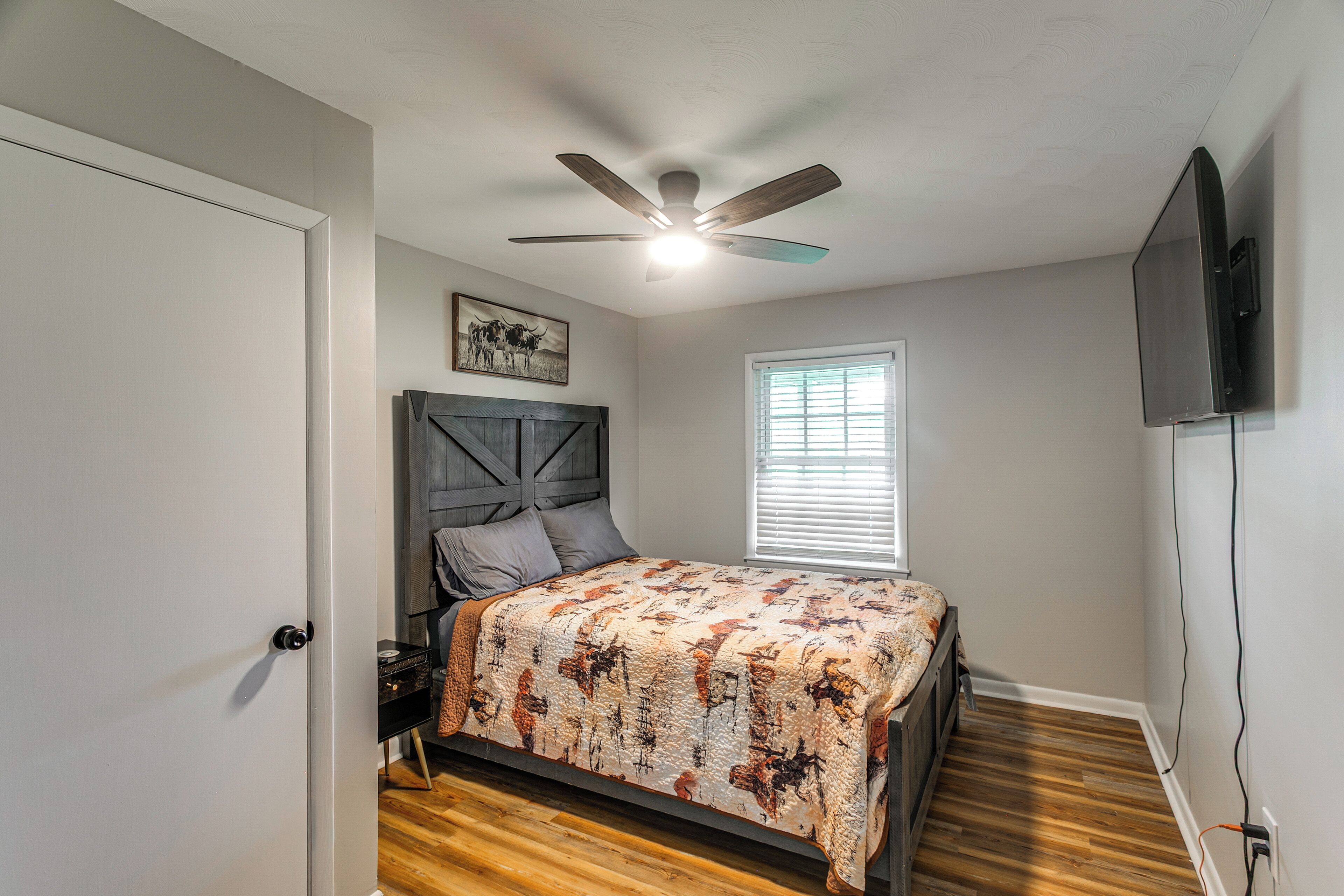 Une chambre à coucher comprenant un lit, un ventilateur de plafond, une fenêtre avec des stores et un téléviseur fixé au mur.