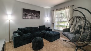 A modern living room with a sectional sofa, a hanging chair, a round ottoman, and a cityscape painting on the wall. - Swim, Golf & Park-Hop! Family Home in Davenport (Davenport)