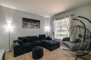 A modern living room with a sectional sofa, a hanging chair, a round ottoman, and a cityscape painting on the wall. - Swim, Golf & Park-Hop! Family Home in Davenport (Davenport)
