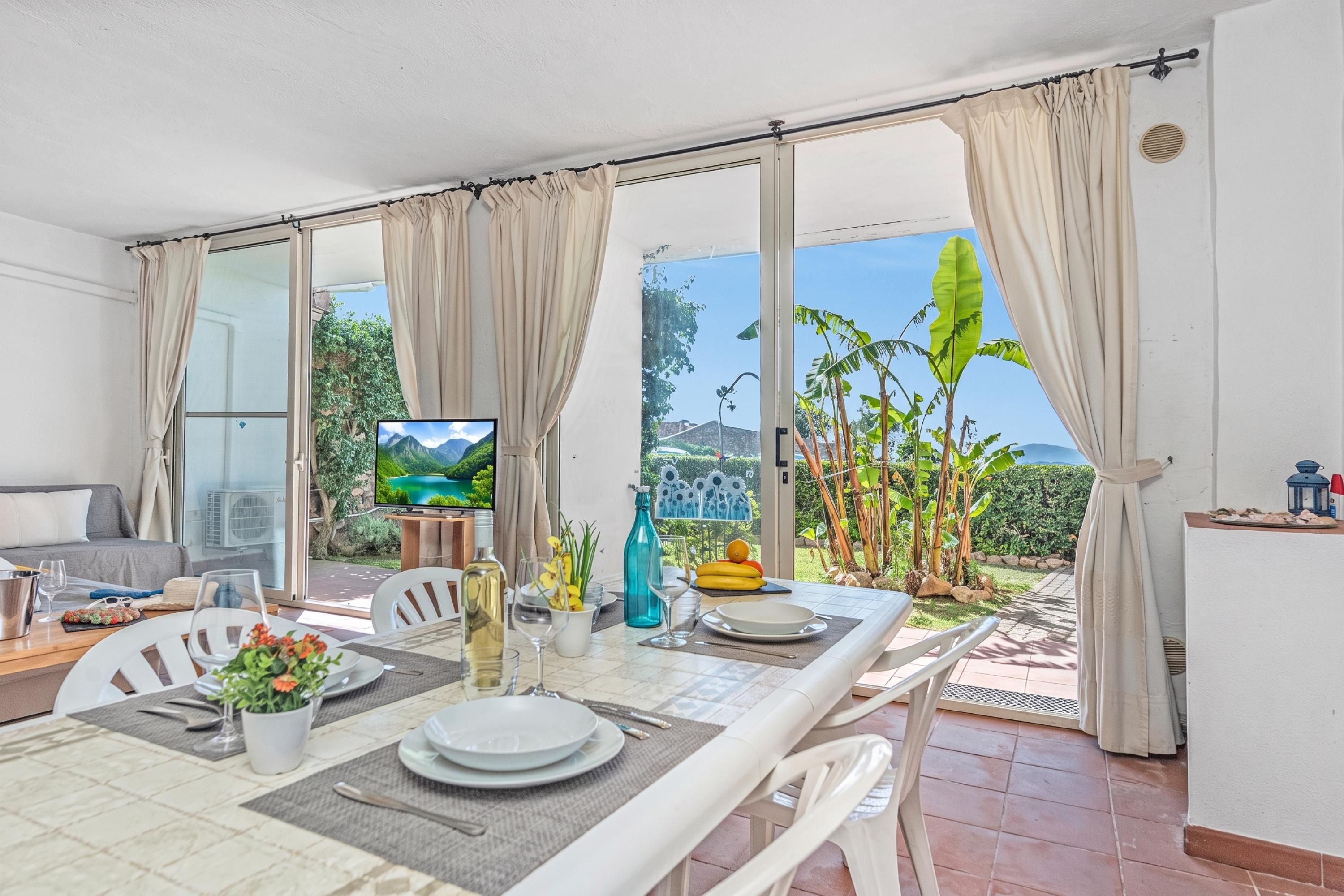 Espaço de refeições com mesa posta para dois, televisão e vista para um jardim tropical.