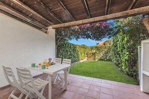 Um pátio com mesa e cadeiras, piso de azulejos e vista para um jardim com cerca de madeira e vegetação.